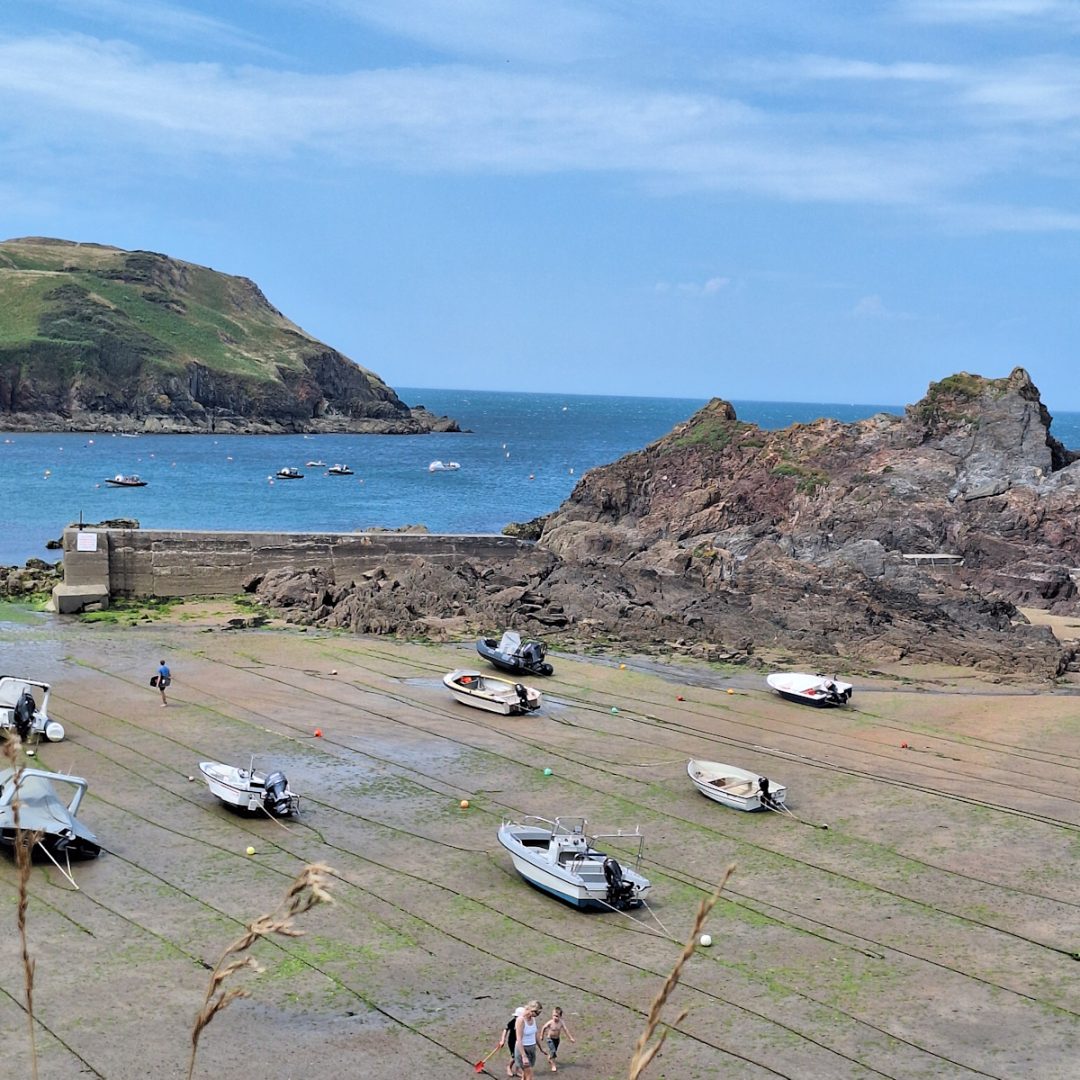 a bay between rocky headlands. The tide is out and several small boats are lying on the beach thethered by long lines. Blue sky with wispy white clouds.